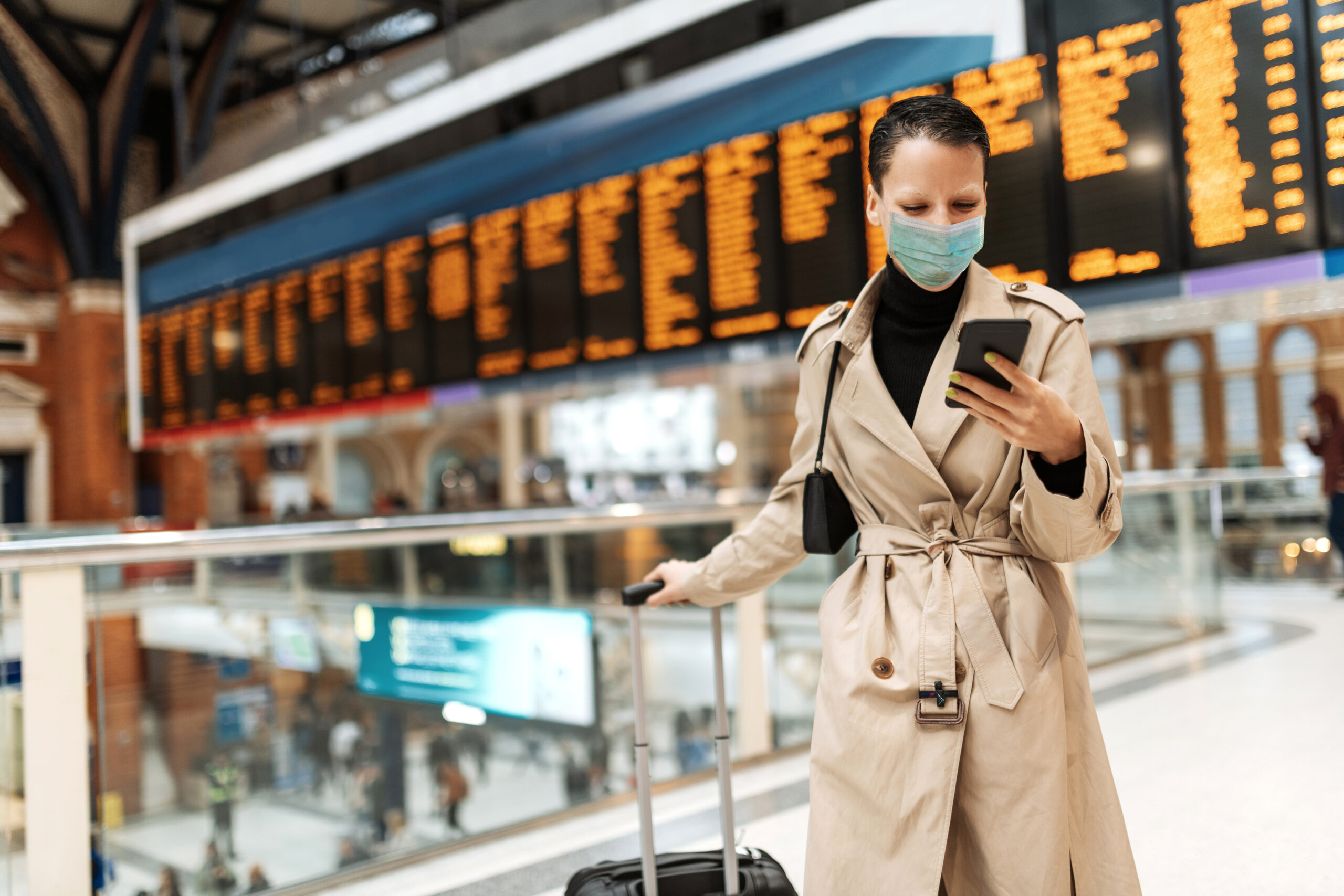Essential travels during lockdown - woman with face mask checking in online while waiting near arrival departure board People around the world wearing face masks to protect themselves and others during Coronavirus pandemic