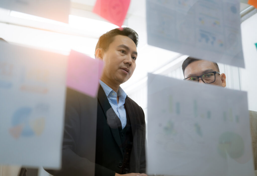 Two men in business attire look at papers and sticky notes on a glass wall, appearing focused during a discussion or brainstorming session in an office setting.