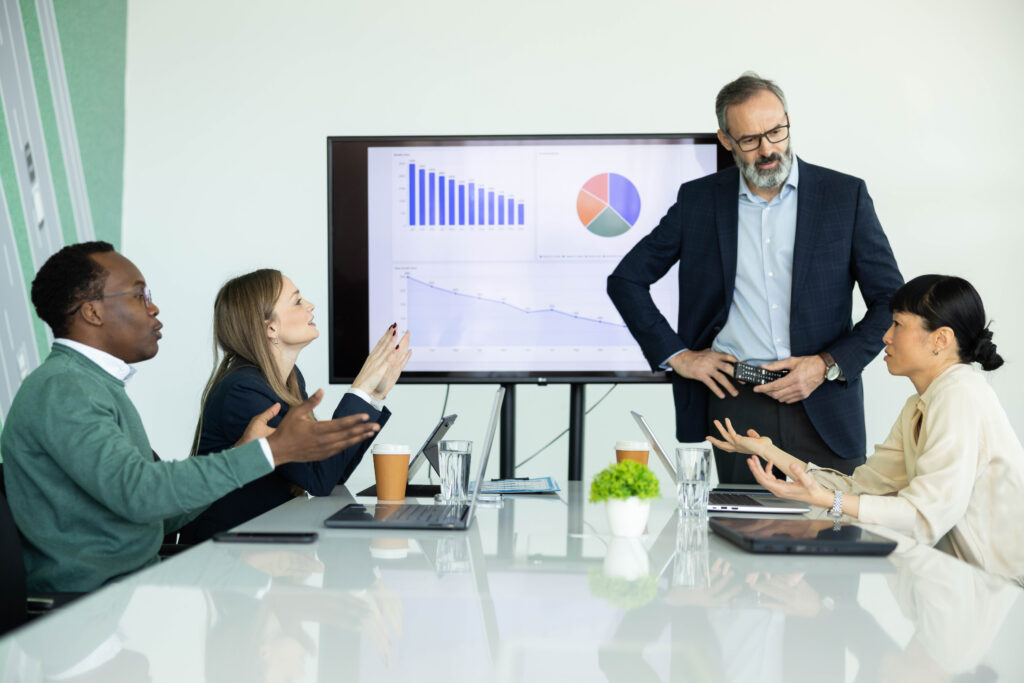 Four people sit around a conference table, having an animated discussion about why AI alone doesn’t move EBIT. A man stands in front of a screen displaying charts and graphs, highlighting the importance of strategic implementation beyond technology. Laptops, papers, and coffee cups are scattered across the table as ideas flow energetically among the team.